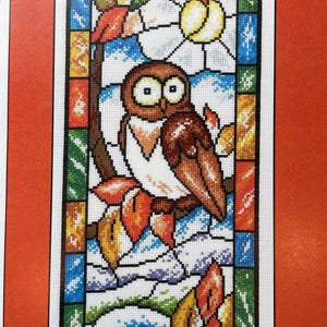 May include: Cross-stitch pattern of an owl perched on a branch, set against a stained-glass window design. The owl has brown and white feathers, with large eyes. The background features a sun, leaves, and a colorful border.