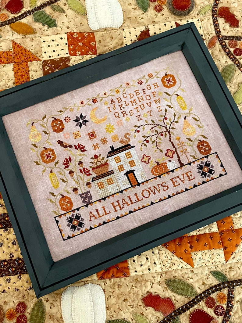 Puede incluir: Un bordado a punto de cruz enmarcado con fondo beige que representa una casa, un &aacute;rbol y calabazas. El texto "ALL HALLOWS EVE" est&aacute; bordado debajo de la casa. El alfabeto est&aacute; bordado en la esquina superior derecha.