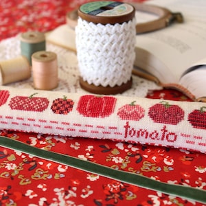 May include: A white pillow with red embroidered tomatoes and the word "tomato" stitched on the bottom. The pillow is on a red and white floral fabric.