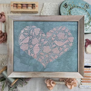 May include: Framed cross-stitch art featuring a heart design filled with pink birds, flowers, and foliage on a teal background. The wooden frame surrounds the artwork. A box of darning and mending cotton is visible above the frame.