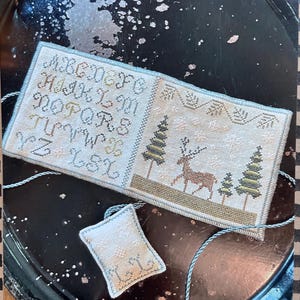 May include: A cross-stitch sampler with the alphabet and a deer scene. The sampler is in a book-like format, with a small, matching tag. The color palette includes beige, green, and brown, with blue accents. The background is a dark, textured surface.