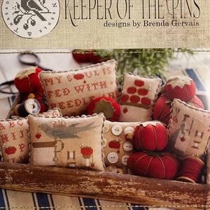 May include: A collection of handmade pincushions in a wooden box. The pincushions are decorated with cross-stitch embroidery and feature various themes, including a bird, a heart, and a needle. The text "KEEPER OF THE PINS" and "designs by Brenda Gervais" is visible on the top of the box.