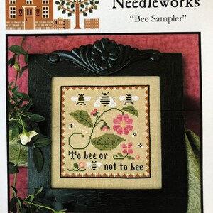 May include: A framed cross-stitch sampler featuring a bee theme. The sampler depicts a floral design with bees and the text "To bee or not to bee". The frame is black with an ornate design.