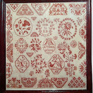 May include: A framed sampler featuring various red cross-stitch designs on a cream-colored background. The sampler includes floral motifs, geometric patterns, and text elements such as "OD to LB" and "A token of love". The frame is a dark brown color.