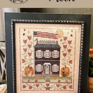 May include: A framed cross-stitch design titled "Harvest Moon" by Annie Beez Folk Art. The design features a brown house with a white picket fence, a pumpkin, and a cat. The house has a red, white, and blue flag hanging from the roof.