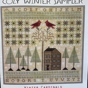 May include: Cross stitch pattern for a winter sampler featuring two red cardinals perched on a tree, a small house, and a snowy forest. The pattern includes the alphabet and the text "Cozy Winter Sampler" and "Winter Cardinals".