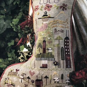 May include: A beige Christmas stocking with a red trim and a colorful embroidered design. The stocking is personalized with the name "Lila" and features a whimsical scene with a house, trees, and animals.