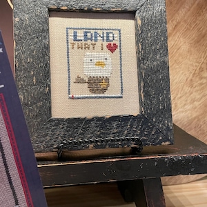 May include: Framed cross-stitch artwork with a patriotic theme. The artwork features an eagle with the text "LAND THAT I" and a heart. The frame is made of distressed black wood, and the piece is displayed on a black stand.