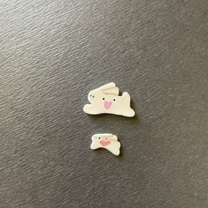 May include: Two white ceramic bunny figurines with pink hearts on their chests. The larger bunny is lying down and the smaller bunny is standing up.