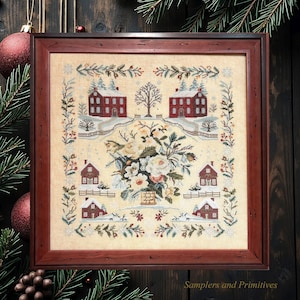 May include: A square cross-stitch embroidery in a dark wood frame featuring a central floral arrangement surrounded by six red houses in a snowy landscape, with a decorative border of pine branches and berries on a light beige fabric background.