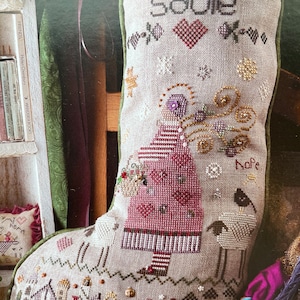 May include: A beige stocking with the name "Sadie" embroidered on it. The stocking features a girl in a red dress holding a basket of flowers, a sheep, and a heart with the word "Hope" embroidered on it.