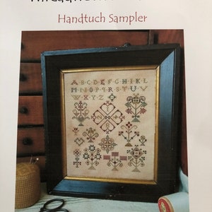 May include: A framed cross-stitch sampler with a cream background and colorful embroidery. The sampler features the alphabet, numbers, and various floral designs. The text "Threadwork Primitives Handtuch Sampler" is at the top of the image. The text "Original Designs by Nan Lewis ~ Threadwork Primitives 2020" is at the bottom of the image.