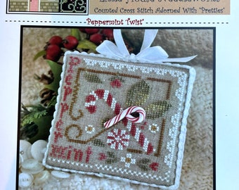 May include: A cross-stitch design titled "Peppermint Twist" by Little House Needleworks. The design features a candy cane, snowflakes, and the word "Peppermint" in red. The piece is framed with a white border and a white ribbon.