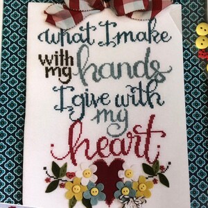 Op de afbeelding: Een witte kruissteek borduurwerk met de tekst "What I make with my hands I give with my heart." Het borduurwerk is ingelijst en bevat een rood hart, bloemen en knopen.