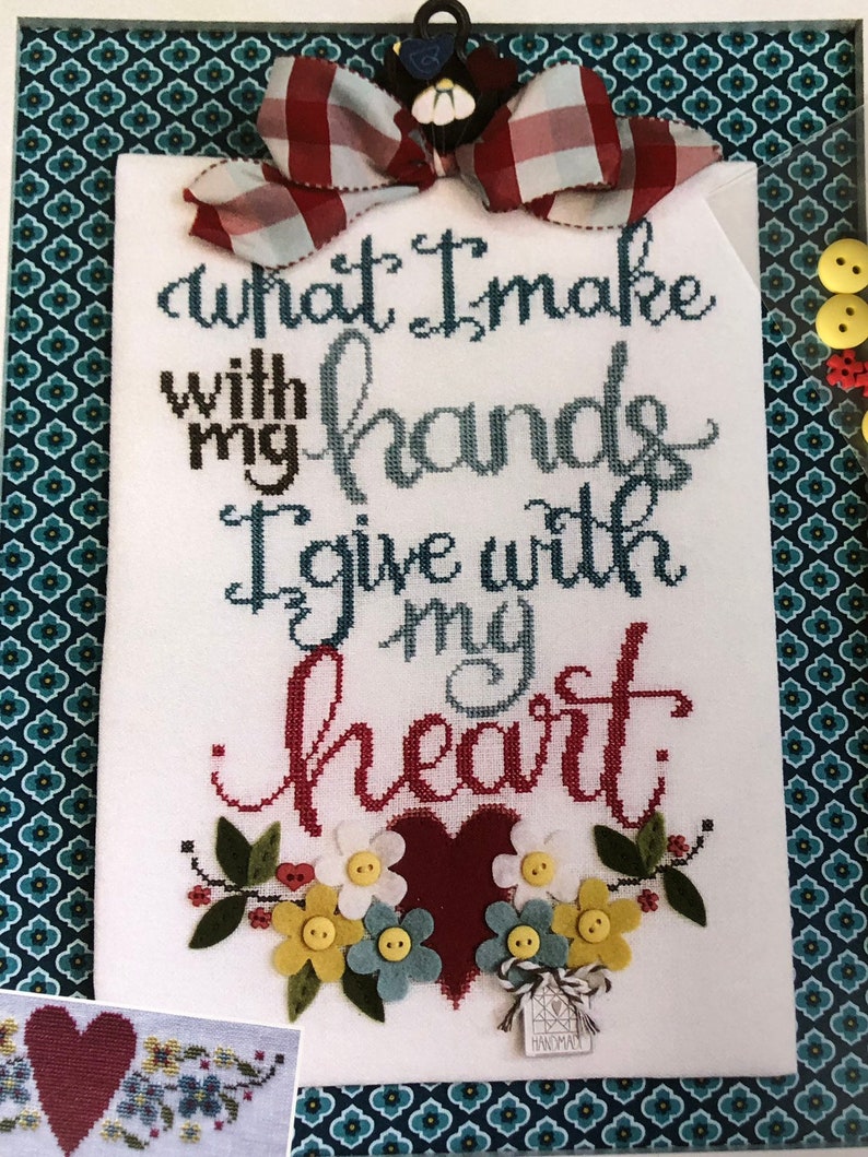 Op de afbeelding: Een witte kruissteek borduurwerk met de tekst "What I make with my hands I give with my heart." Het borduurwerk is ingelijst en bevat een rood hart, bloemen en knopen.