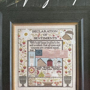 May include: A cross-stitch sampler with a black frame and a beige background. The sampler features the text "Declaration of Sentiments" and the quote "We hold these truths to be self-evident that all men and women are created equal..." with the years 1848 and 1920. The sampler also includes the names Lucretia Mott, Elizabeth Cady Stanton, and Mary Ann McClintock.