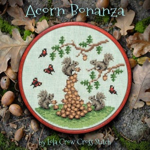 May include: A cross-stitch embroidery hoop with a red frame features a design titled "Acorn Bonanza." The artwork depicts squirrels, acorns, butterflies, and tree branches. The text "by Lola Crow Cross Stitch" is at the bottom.