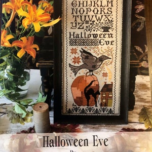 May include: A framed cross-stitch design titled "Halloween Eve" by Blackbird Designs. The design features a black cat, a bird, a pumpkin, and a small house. The alphabet is stitched above the design.