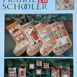 May include: A book cover titled "The Prairie Schooler" featuring cross-stitch patterns for Christmas stockings and ornaments. The stockings depict houses, Santa Claus, and other holiday motifs. The color palette includes beige, red, and green.