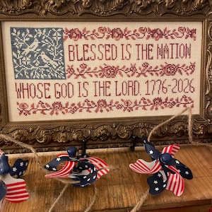 May include: Framed cross-stitch artwork with the text "BLESSED IS THE NATION WHOSE GOD IS THE LORD. 1776-2026" in red. The frame is ornate gold. Patriotic pinwheel ornaments with red, white, and blue ribbons hang below.