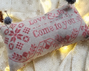 May include: A rectangular, cross-stitched pillow with the words "Love & Joy Come to You" in red thread. The pillow is decorated with floral and snowflake patterns. The background features a patterned fabric and fairy lights. The text "Love & Joy" is also displayed.