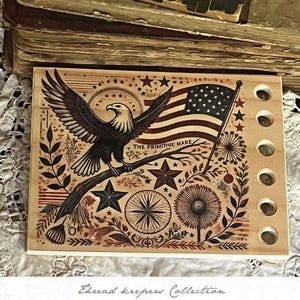 May include: Wooden thread keeper featuring an eagle perched on a branch, an American flag, and star designs. The text "EAGLE THREAD KEEP" and "2025 SUMMER COLLECTION BY THE PRIMITIVE HARE" are visible. The thread keeper has six holes.