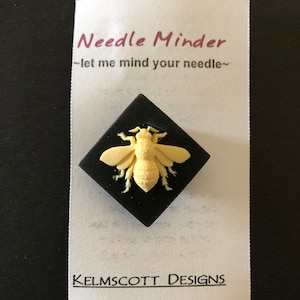 May include: A black square needle minder with a white 3D bee design. The needle minder has the text "Needle Minder" and "~let me mind your needle~" on a white card. The card also has the text "KELMSCOTT DESIGNS".