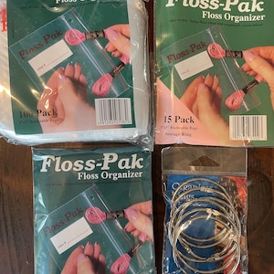May include: Four packages of floss organizer supplies. Three packages are labeled "Floss-Pak" and contain clear reclosable bags with pink floss. The fourth package contains silver storage rings.