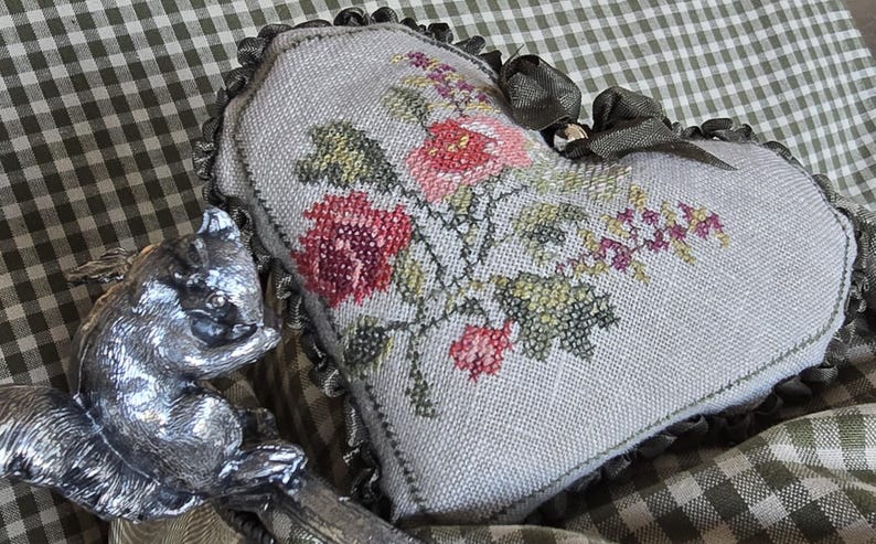 May include: A heart-shaped pillow with a floral embroidery design. The pillow is made of a light-colored fabric and features a delicate floral pattern in shades of pink, red, and green. The pillow is accented with a brown ribbon.
