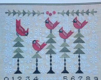 May include: Cross-stitch sampler with red birds perched on green trees, snowflakes, and the word "SAMPLER" at the top. Numbers 0-9 are at the bottom. The design is on a beige background.