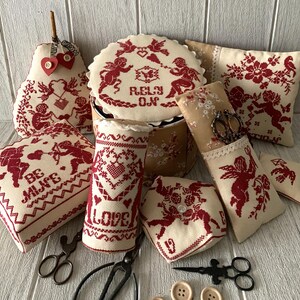 May include: A collection of handmade Valentine's Day themed needlepoint pillows and a round box with a lid. The pillows feature red and white embroidery with cherubs, hearts, and the words "Be Mine", "Love", and "Rely On".