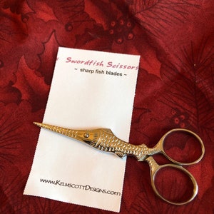 May include: Silver-toned swordfish-shaped scissors with sharp fish blades. The scissors are on a white card with the text "Swordfish Scissors - sharp fish blades - www.KELMSCOTTDESIGNS.com".