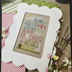 May include: A white frame with a scalloped edge surrounds a cross-stitch design featuring a whimsical scene of a raincloud, raindrops, an umbrella, and chicks with the text "Rain drop kisses fill your wishes". The frame is on a pink and white gingham background with a green floral design.