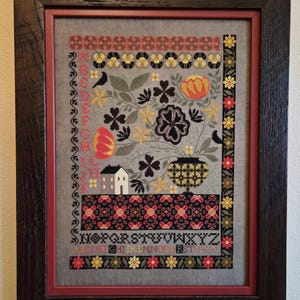 May include: Framed cross-stitch artwork featuring floral and geometric patterns in red, yellow, black, and gray. The design includes flowers, birds, a house, and the alphabet. The artwork is framed in a dark wood frame with a red inner border.