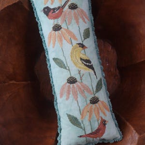 May include: A rectangular, light blue decorative pillow with a stitched design of birds and flowers. The pillow features a teal fringe and a floral and bird pattern in shades of orange, green, and red. The pillow is placed in a wooden bowl.
