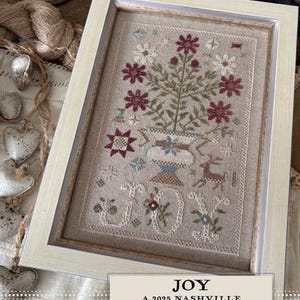 May include: A framed cross-stitch design with a floral motif in shades of red, white, and green. The design features a vase with flowers and the word "JOY" in a stylized font. The frame is white with a light brown mat. The text below the design reads "JOY A 2025 NASHVILLE EXCLUSIVE - by Shakespeare's Peddler - "
