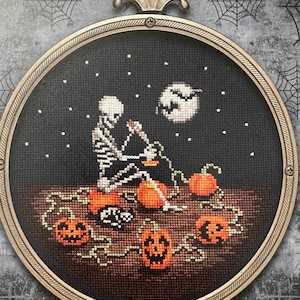 May include: A cross-stitch design featuring a skeleton with a lantern, surrounded by orange pumpkins, under a starry night sky with a full moon. The artwork is framed in a round, ornate, bronze-colored frame, perfect for Halloween decor.