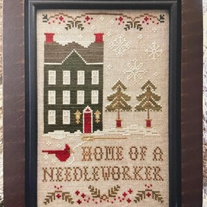 May include: Cross-stitch artwork in a dark wooden frame. The design features a house with a green roof and red chimney, two pine trees, and the text "HOME OF A NEEDLEWORKER". Snowflakes and berries decorate the top and bottom.