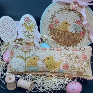 May include: A cross-stitch pillow with a floral design and two yellow chicks. The pillow is surrounded by wooden spools, pink eggs, and straw. The text on the pillow reads "Change is Coming".