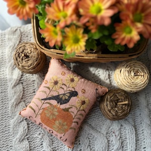 Peut inclure: Un coussin rose avec un motif de point de croix représentant un oiseau noir perché sur une citrouille avec des tournesols. Le coussin est posé sur une couverture en tricot blanc à câble avec des boules tissées brunes et beiges.