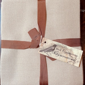May include: A square piece of light beige fabric with a visible woven texture, folded and tied with a brown ribbon. A rectangular paper tag is attached, featuring a bird illustration and the text 32ct Cappuccino Nikyscreations.