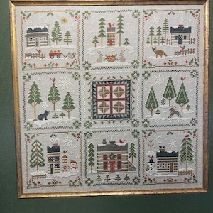 May include: A framed cross-stitch sampler with a winter theme. The sampler features nine different scenes of houses, trees, and snow. The houses are all different colors and styles, and the trees are all green. The snow is white and fluffy.