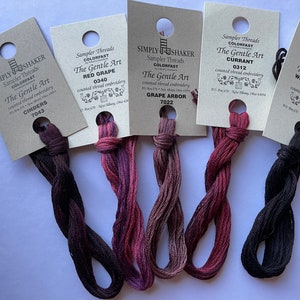 May include: A collection of six skeins of The Gentle Art sampler threads in various shades of purple, red, and black. Each skein is attached to a small, brown paper tag with the thread color name and number printed on it. The threads are arranged on a white surface.