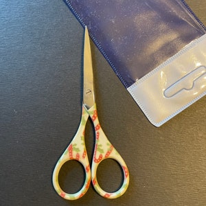 May include: A pair of small, silver-colored scissors with a green, red, and white patterned handle. The scissors are on a dark gray surface.