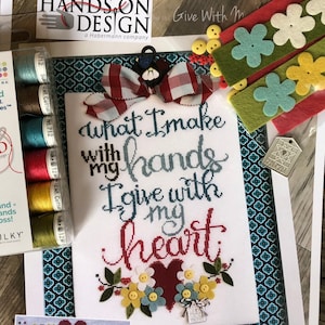 Op de afbeelding: Borduurpatroonset met een rood hart en de tekst "what I make with my hands I give with my heart". De set bevat borduurgaren, vilt, knopen en een klein label.