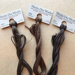 May include: Three spools of Weeks Dye Works floss in brown, dark brown, and black shades. Each spool is labeled with the brand name, "Weeks Dye Works", the collection name, "Floss Collection", and the color name and number. The colors are Grape Vine (1273), Chestnut (1269), and Tatanka (1272).