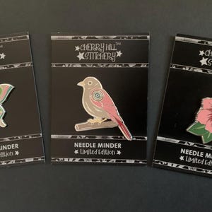 May include: Three enamel pins featuring a butterfly, bird, and flower design. Each pin includes the text "Cherry Hill Stitchery" and "Needle Minder Limited Edition".