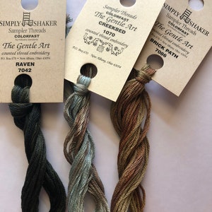 May include: Three skeins of embroidery floss in shades of green, blue, and brown. The floss is labeled with the brand name "Simply Shaker" and the color names "Raven 7042", "Creekbed 1070", and "Brick Path 7086".