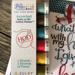 Op de afbeelding: Een verpakking met zes verschillende Sulky katoenen borduurgaren in 12-draads gewicht in verschillende kleuren, waaronder teal, bruin, rood, geel en groen. De verpakking is gelabeld met "Cotton Petites" en "Hands On Design".