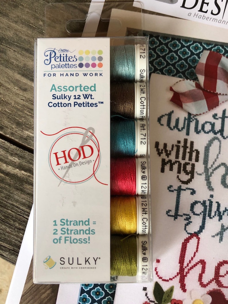 Op de afbeelding: Een verpakking met zes verschillende Sulky katoenen borduurgaren in 12-draads gewicht in verschillende kleuren, waaronder teal, bruin, rood, geel en groen. De verpakking is gelabeld met "Cotton Petites" en "Hands On Design".
